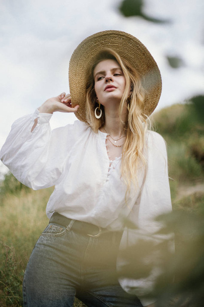 stylish woman in straw hat and white linen blouse and jeans posing on meadow at countryside - Photo, Image