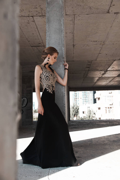Glamour fashion model in long evening dress posing in abadoned place. young beautiful woman in luxury black dress - Foto, Imagen