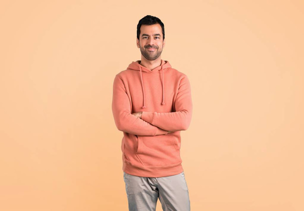 Mann im rosafarbenen Sweatshirt, der die Arme frontal verschränkt hält. Selbstbewusster Ausdruck auf ockerfarbenem Hintergrund - Foto, Bild