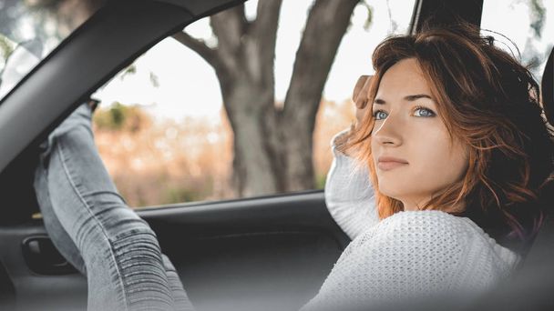 girl with red hair in a white sweater and gray jeans is sitting in the car in the front seat with her legs out the window - Foto, Imagem