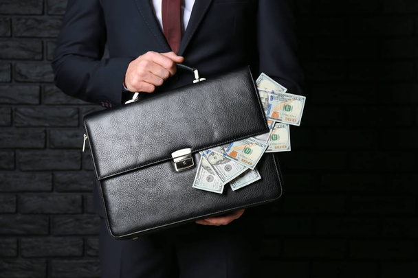 Hombre de negocios sosteniendo maletín con billetes de dólar sobre fondo oscuro. Concepto de corrupción
 - Foto, Imagen
