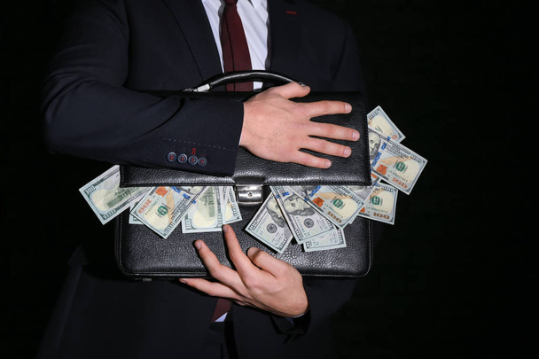 Businessman holding briefcase with dollar banknotes on dark background. Corruption concept - Photo, Image