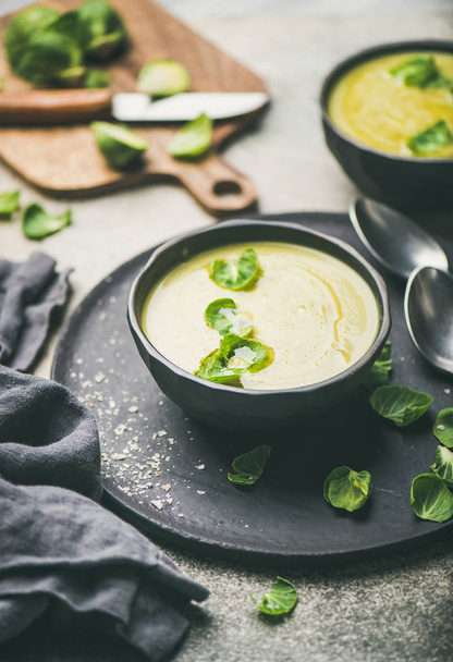 Seasonal brussels sprouts vegetable cream soup in black bowl and fresh green brussel sprouts over grey concrete background, vertical composition. Vegan, vegetarian, healthy, dieting food concept - Fotografie, Obrázek