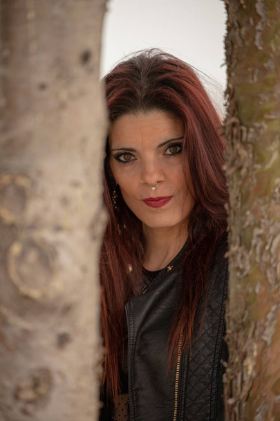 Posed redheaded woman with piercing in the nose and dressed in black jacket next to a tree - Photo, Image