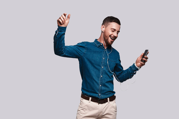 komea nuori mies kuuntelee musiikkia ja hymyilee seistessään harmaata taustaa vasten matkapuhelin kädessä
 - Valokuva, kuva