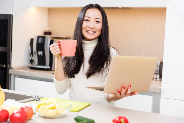Image of young brunette with cup in hand at table with laptop in kitchen - Foto, Imagen