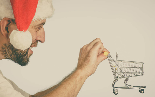 Man wearing santa claus hat holding empty shopping basket cart. Xmas, seasonal sales, boxing day concept. - Fotografie, Obrázek