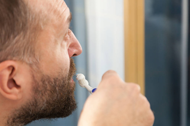 Adult man brushing his teeth looking at his bathroom mirror during morning hygiene routine. - Фото, изображение