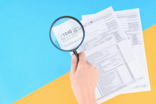 Ausgeschnittene Ansicht einer Frau mit Lupe über Steuerformulare auf blauem und gelbem Hintergrund - Foto, Bild
