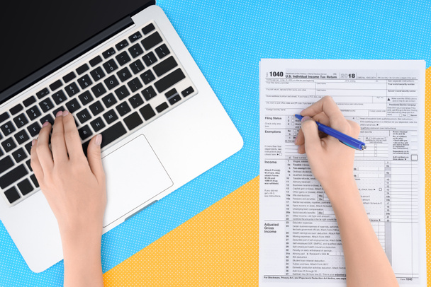 bijgesneden beeld van de vrouw te typen op de laptop en vullen belastingformulier op blauwe en gele achtergrond - Foto, afbeelding