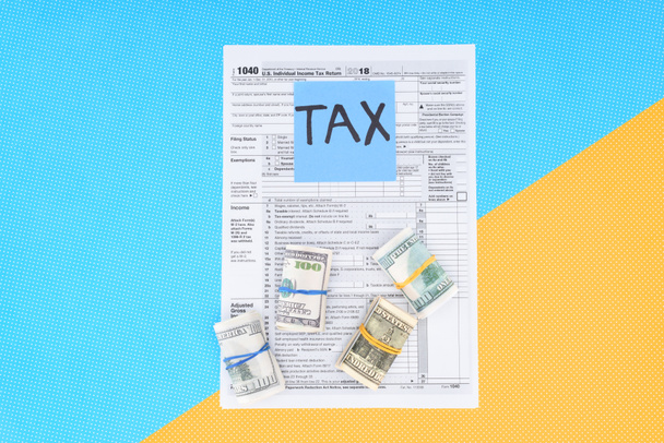 top view of tax form, money rolls and card with 'tax' word on blue and yellow background - Fotó, kép