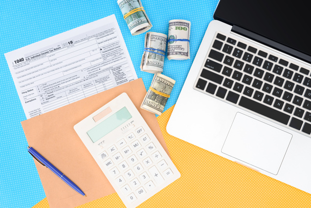 bovenaanzicht van belastingformulier, geld rollen en laptop op blauwe en gele achtergrond - Foto, afbeelding