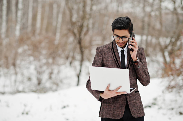 Stylish indian student man in suit and glasses posed at winter day outdoor with laptop at hands, speaking at phone. - Фото, изображение