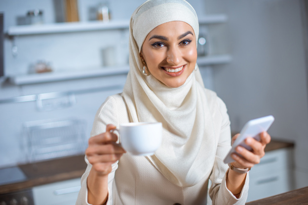 smiling young muslim woman holding cup of coffee and smartphone at home - Foto, Imagem
