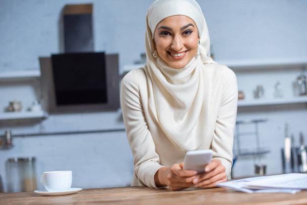 mooie jonge moslimvrouw gebruikend smartphone en lachend op camera  - Foto, afbeelding