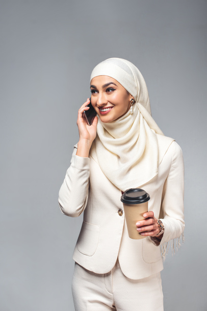 mujer musulmana sonriente hablando por teléfono inteligente y sosteniendo café para ir aislado en gris
  - Foto, imagen