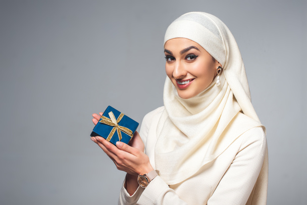happy young young muslim woman holding present and smiling at camera isolated on grey  - Zdjęcie, obraz
