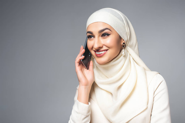 mooie gelukkig jonge moslimvrouw praten door smartphone en op zoek weg geïsoleerd op grijs - Foto, afbeelding