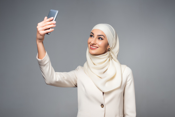 hermosa mujer musulmana sonriente tomando selfie con teléfono inteligente aislado en gris
   - Foto, imagen