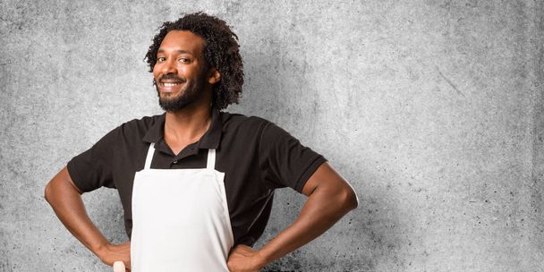 Handsome african american baker with hands on hips, standing, relaxed and smiling, very positive and cheerful - Foto, immagini