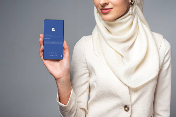 cropped shot of smiling muslim woman holding smartphone with facebook app isolated on grey  - 写真・画像