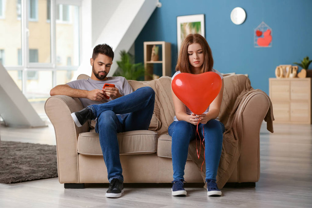 Mulher jovem triste segurando balão em forma de coração perto homem indiferente brincando com telefone celular em casa. Problemas de relacionamento
 - Foto, Imagem