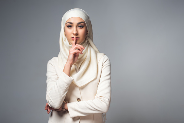 mladá muslimka stojí s prstem na rtech a při pohledu na fotoaparát izolované Grey  - Fotografie, Obrázek