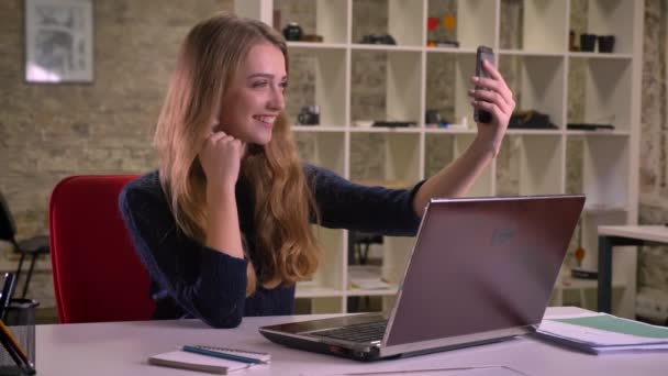 Portrait of blonde caucasian businesswoman in front of laptop in office making selfie-photos using smartphone. - Filmati, video