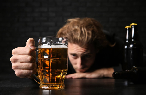 Humalainen nuori mies oluen kanssa pöydässä lähellä mustaa tiiliseinää. Alkoholismin käsite
 - Valokuva, kuva