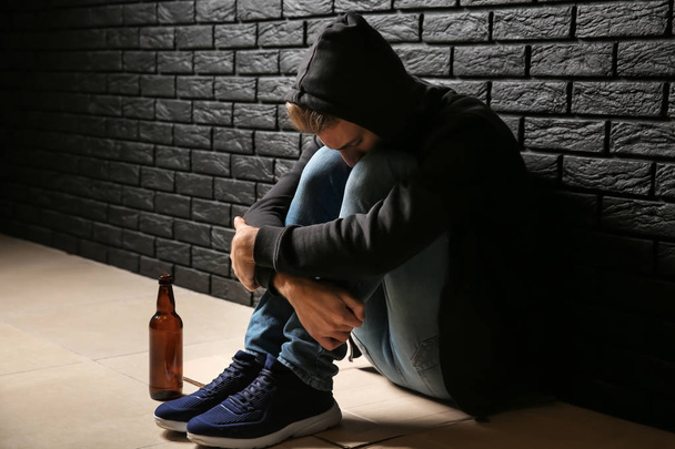 Przygnębiony młody człowiek z butelką piwa, siedząc na podłodze w pobliżu czarny ceglany mur. Pojęcie alkoholizmu - Zdjęcie, obraz