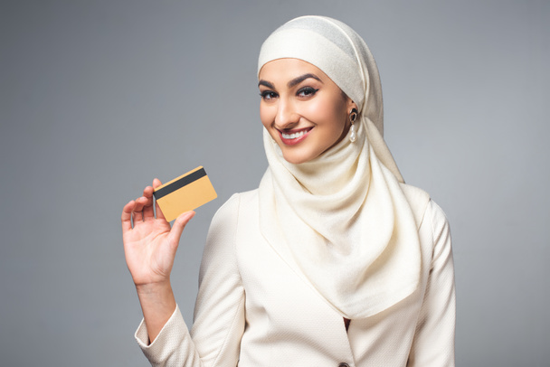 молода мусульманка тримає кредитну картку і посміхається на камеру ізольовано на сірому
  - Фото, зображення