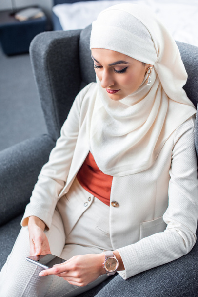 beautiful young muslim woman sitting in armchair and using smartphone  - Foto, afbeelding