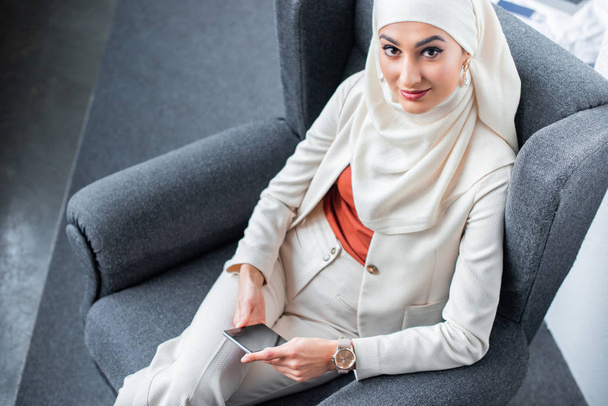 high angle view of beautiful muslim woman using smartphone and smiling at camera  - Фото, изображение