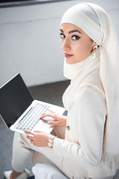 attractive muslim woman using laptop with blank screen and looking at camera  - Foto, afbeelding