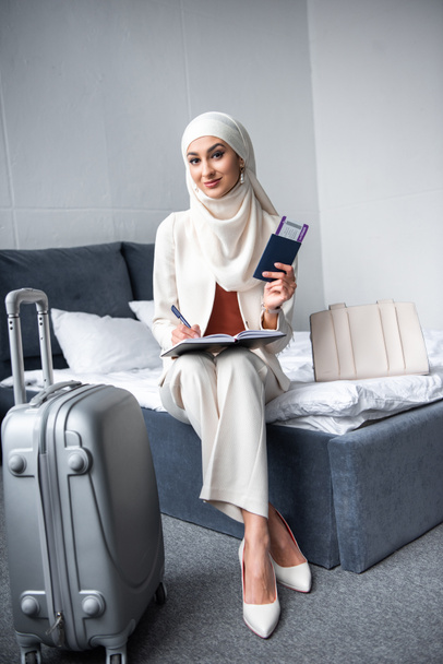 beautiful muslim woman holding passport with boarding pass and smiling at camera in bedroom   - Fotó, kép