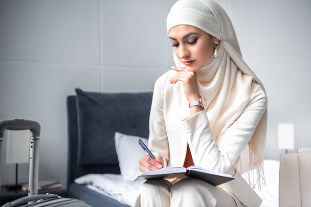 Yatağın üstüne oturan ve not defterinde yazılı düşünceli Müslüman kadın - Fotoğraf, Görsel