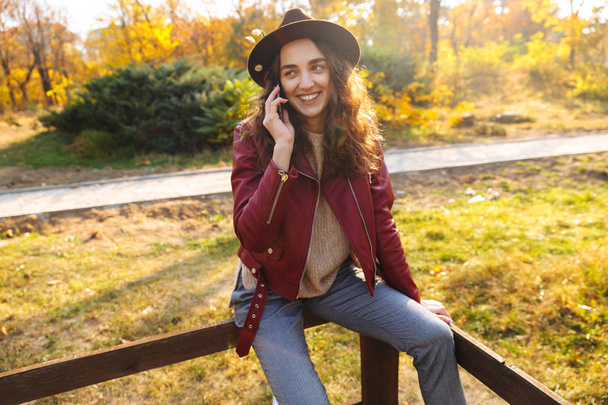 Beautiful young woman wearing hat at the park in autumn, talking on mobile phone - Zdjęcie, obraz