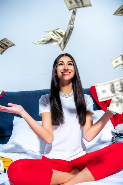 cheerful young brunette woman sitting on bed and throwing dollar banknotes - Foto, afbeelding