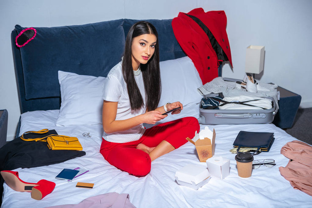 Hochwinkelaufnahme des schönen brünetten Mädchens mit Smartphone und Blick auf die Kamera, während es mit dem Koffer vor der Reise im Bett sitzt - Foto, Bild