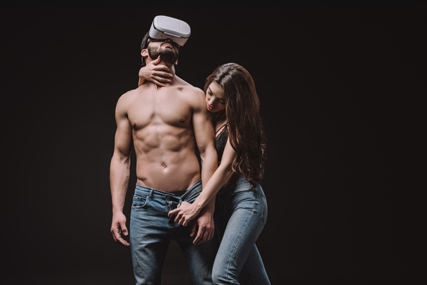 beautiful girl undressing shirtless man in vr headset isolated on black - Foto, imagen