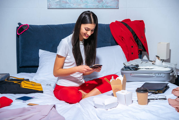 mooie lachende brunette meisje met behulp van smartphone zittend op bed met koffer voor reis - Foto, afbeelding