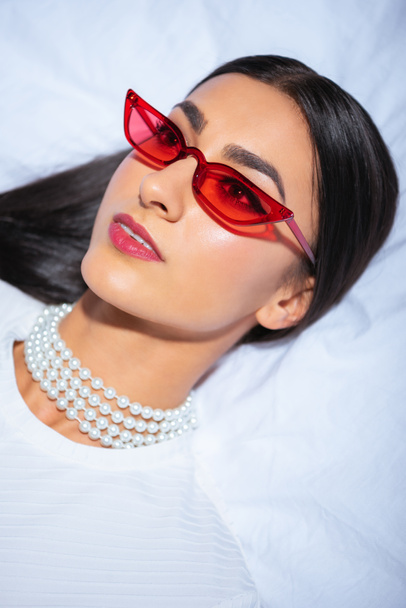 elegant young woman in red eyeglasses lying on bed and looking away - Photo, Image