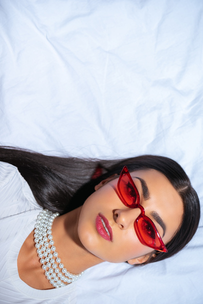 top view of beautiful young woman in red sunglasses lying on bed and looking at camera  - Foto, Imagem