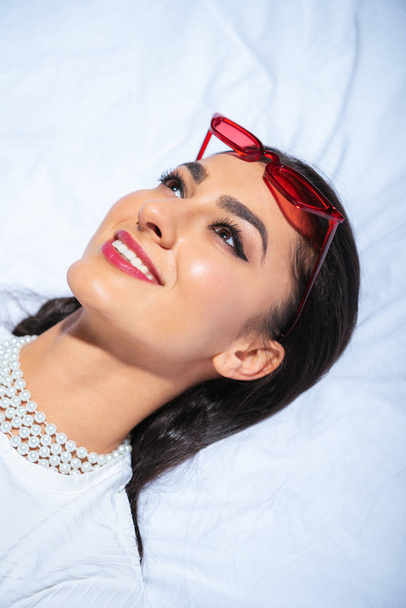 beautiful smiling young brunette woman with red sunglasses on head lying on bed and looking up - 写真・画像