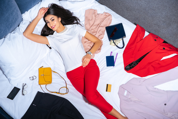 beautiful girl smiling at camera while lying on bed with fashionable clothes, handbags with money, credit card and passport with ticket  - Foto, imagen
