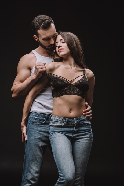 handsome man undressing girl in lace bra isolated on black - Photo, Image
