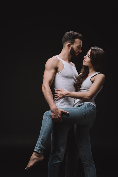 handsome man hugging woman in white singlet and jeans isolated on black - Photo, Image