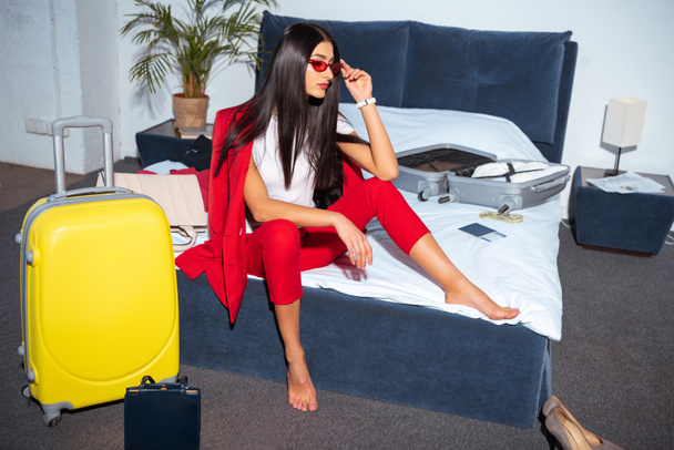 high angle view of beautiful young woman in fashionable red suit and sunglasses sitting on bed and looking away - Foto, Bild