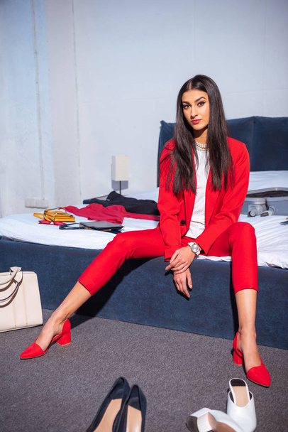 attractive young woman in stylish red suit looking at camera, various fashionable shoes on floor and clothes on bed - Foto, imagen