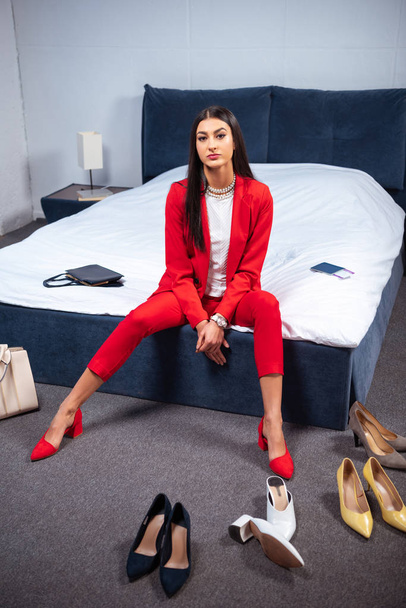 beautiful young woman in stylish red suit sitting on bed and looking at camera, various fashionable shoes on floor    - Фото, изображение